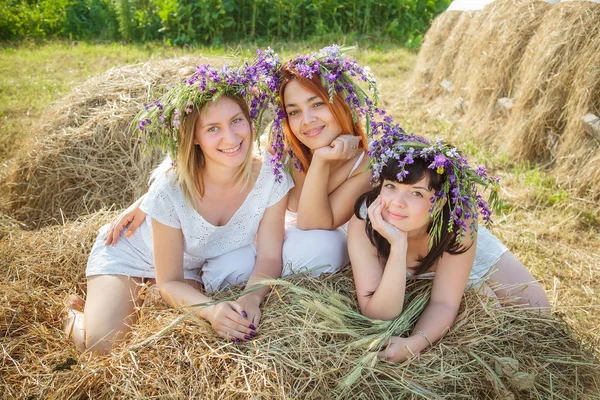 Tre giovani belle ragazze in abiti bianchi e ghirlande di fiori selvatici seduti vicino al pagliaio. Estate nel villaggio — Foto Stock