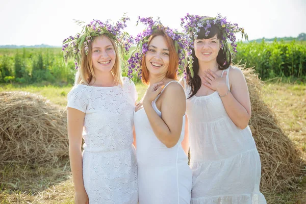 Tre giovani belle ragazze in abiti bianchi e ghirlande di fiori selvatici che soggiornano vicino al pagliaio e ridono. Estate nel villaggio — Foto Stock