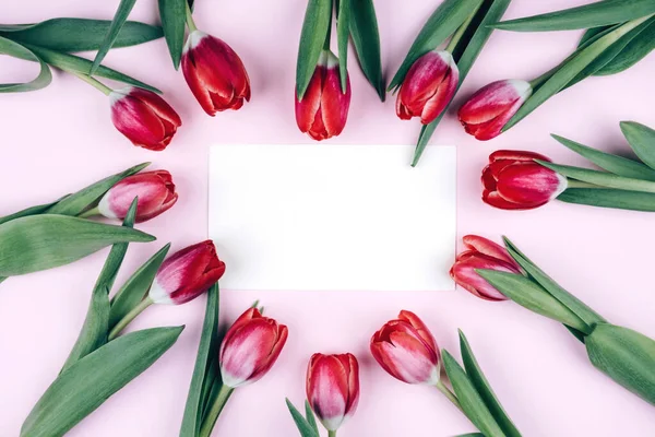 Red tulips are laid out in the heart on a pink background with a postcard.