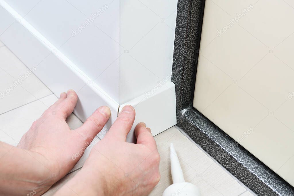 A man with glue glues floor MDF plinth.A man sets up white floor moldings in an apartment.Close view of man installing new skirting board.