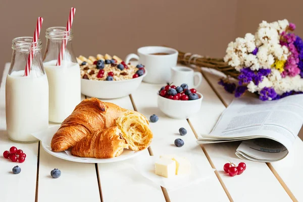 Смачний сніданок з круасанами, вівсяними пластівцями, ягодами та молоком на білому дерев'яному столі — стокове фото