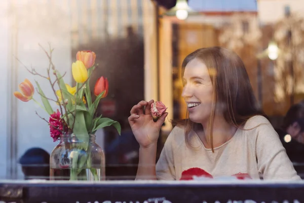 Hermosa mujer joven riendo sentada en la cafetería cerca de la ventana, secando café y comiendo postre —  Fotos de Stock