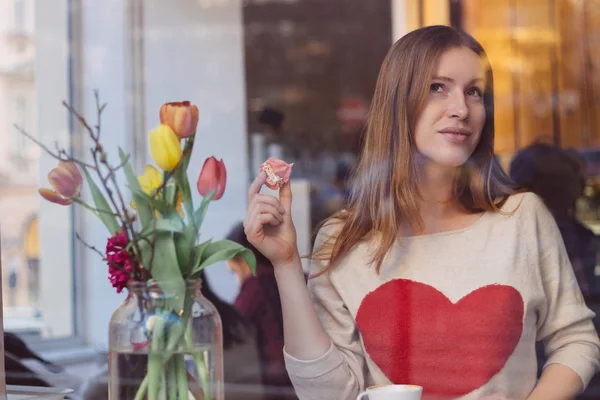 Hermosa joven soñadora sentada en una cafetería cerca de la ventana, tomando café y comiendo postre —  Fotos de Stock