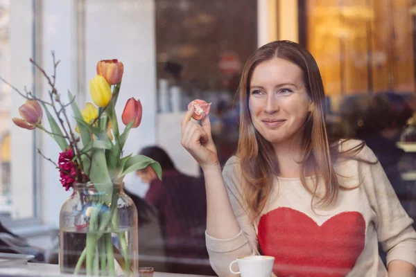 Hermosa joven sonriente sentada en la cafetería cerca de la ventana, tomando café y comiendo postre —  Fotos de Stock