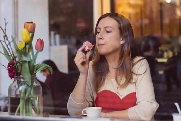 Hermosa joven sentada en la cafetería cerca de la ventana, tomando café y comiendo postre —  Fotos de Stock