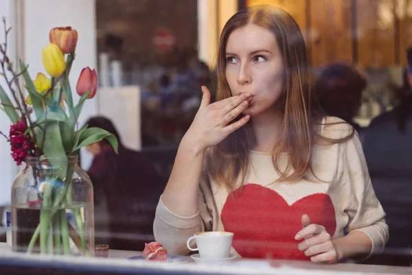 Hermosa joven sentada en la cafetería, mirando por la ventana, tomando café y comiendo postre —  Fotos de Stock