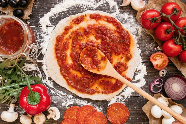 Cocinar pizza. Añadir salsa de tomate fresco a la masa de pizza. Ingredientes de pizza en la mesa de madera vista superior — Foto de Stock