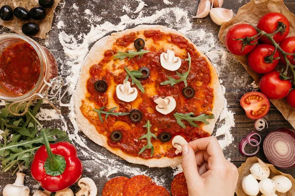 Cocinar pizza. Manos añadiendo ingredientes a la pizza. ingerentes de pizza en la mesa de madera, vista superior — Foto de Stock