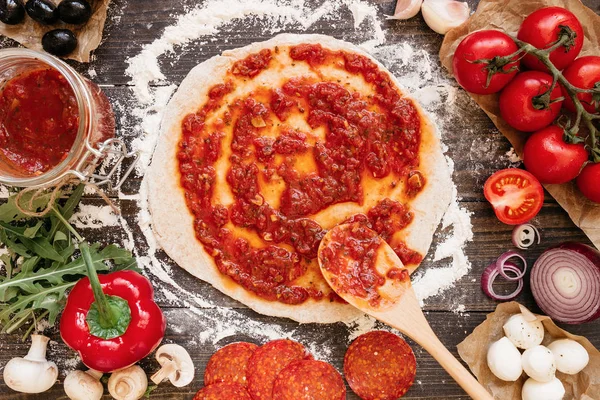 Cocinar pizza. Añadir salsa de tomate fresco a la masa de pizza. Ingredientes de pizza en la mesa de madera vista superior — Foto de Stock