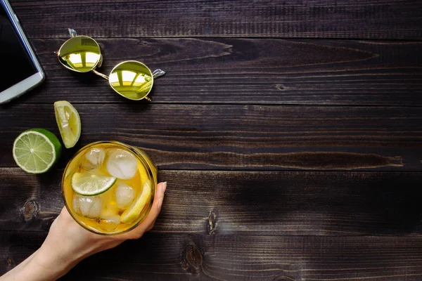 Les mains tenant une boisson glacée. Thé glacé, lunettes de soleil et smartphone sur la table en bois, vue sur le dessus. Avec espace de copie — Photo