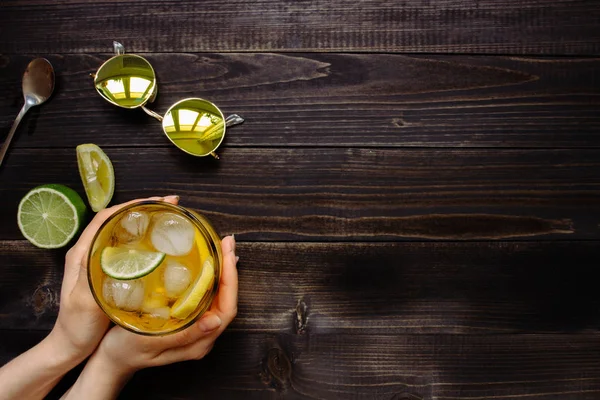 Les mains tenant une boisson glacée. Thé glacé, citron, lime et lunettes de soleil sur la table en bois, vue sur le dessus. Avec espace de copie — Photo