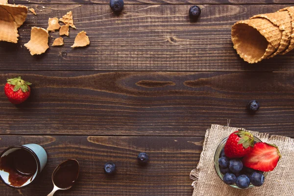 Bayas frescas, cono de gofre y salsa de chocolate en el fondo de madera, vista superior. Fondo de helado con espacio de copia — Foto de Stock