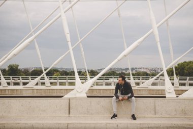 Onun smartphone kullanarak şehrin modern köprüde oturan genç adam