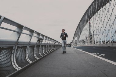 Şehrin modern köprü üzerinde çalışan, smartphone cep telefonu ile müzik dinleme genç adam