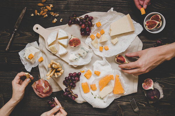 Eating different kinds of cheeses with fruits and snacks on the wooden dark table. Top view
