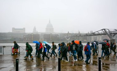 Londra / Birleşik Krallık - 24 Ekim 2019: Londra 'da şiddetli yağmur. Milenyum köprüsü ve St. Paul Katedrali 'nin kubbesi ile Thames nehrinin güney kıyısından insanlar geçiyor.