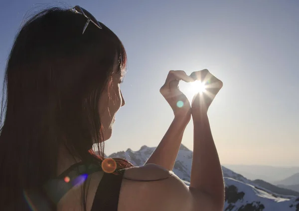 Το κορίτσι διπλωμένα τα χέρια της σε σχήμα καρδιάς μέσα από τις ακτίνες του ήλιου φόντο τα βουνά — Φωτογραφία Αρχείου