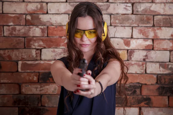 beautiful, young girl with a gun in hands, wearing glasses and headphones against a brick wall background