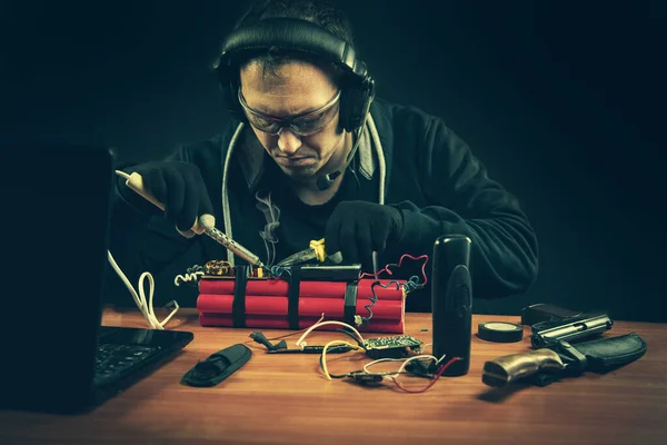 蒙面恐怖分子手拿着焊铁在室内准备爆炸装置 放在桌子上 用手机 手提电脑制造的雷管 — 图库照片