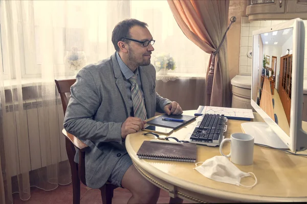 Sitting at the kitchen table, a young man in a business suit and shorts holds works extended meeting with a business partner on social networks,