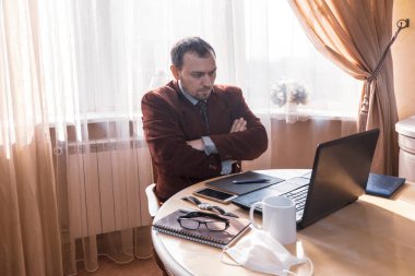 Uzaktan çalışma, mutfak masasında oturan takım elbiseli ve şortlu genç bir adam, iş fikirlerini dikkatle okuyor, kollarını kavuşturuyor.