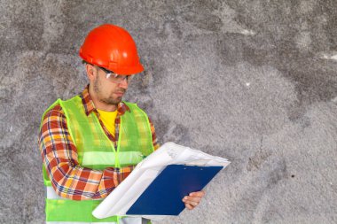 İnşaatçı, ustabaşı, koruyucu giysiler içinde, bir kask, yansıtıcı ceket ve gözlük, beton duvar arka planındaki çizim ve diyagramlara bakıyor..