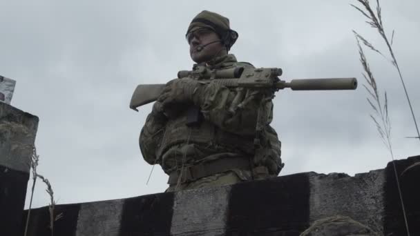 Militar en un uniforme del ejército y sombrero de pie en un campo sobre un fondo de cielo y hierba. Un soldado con un arma cerca del refugio — Vídeo de stock