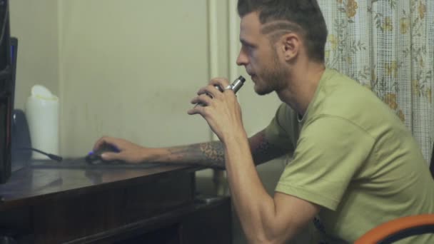 Молодой человек садится за стол и курит электронную сигарету, разрешает парам — стоковое видео