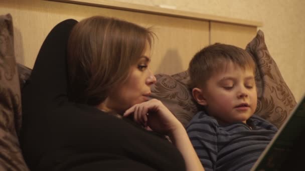 Молода мати разом маленький син читає книгу, що лежить на ліжку в затишній спальні — стокове відео
