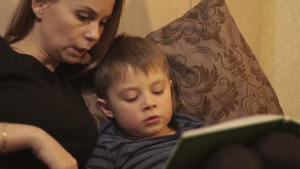 Joven madre juntos pequeño hijo leyendo un libro acostado en la cama en el acogedor dormitorio — Vídeo de stock