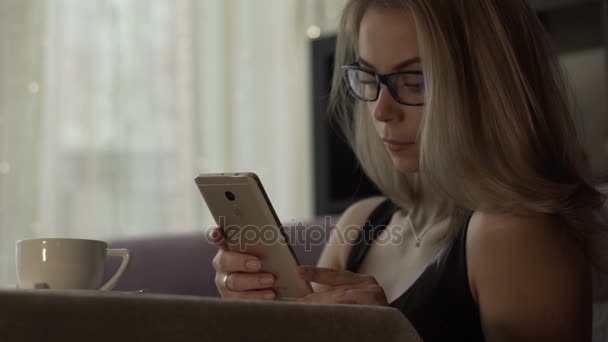 Mujer joven en gafas usando teléfono móvil con taza de café de pie en la mesa — Vídeos de Stock