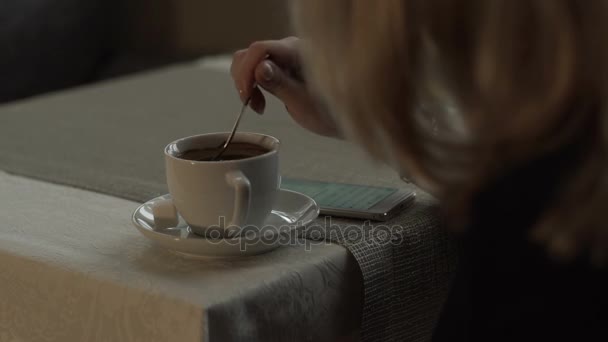 Jovem mulher misturando açúcar na xícara de café na mesa no fundo do telefone móvel — Vídeo de Stock