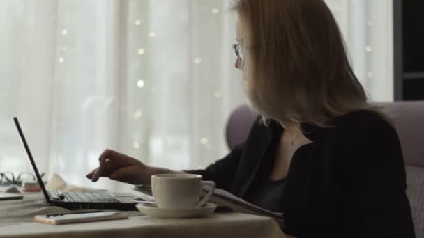Mujer de negocios en gafas de escribir en el teclado portátil sentado en el sofá en la cafetería — Vídeos de Stock