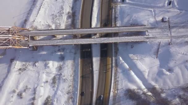 Cars driving on winter highway under suspension railway bridge drone view. Car traffic on snowy road on winter landscape. Aerial view train bridge over highway road. — Stock Video
