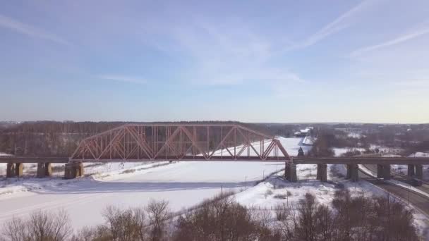 Suspension railway bridge for train traffic over frozen river on winter landscape aerial view. Car traffic on winter highway over train bridge drone view. — Stock Video