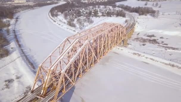 Railway bridge and railway track for train traffic over frozen river on winter landscape drone view. Suspension train bridge through river and car highway aerial view. — Stock Video
