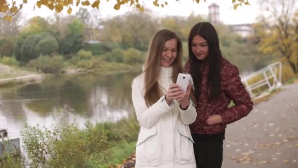 Two beautiful girls are walking in autumn park, watching a photo on the phone — Αρχείο Βίντεο