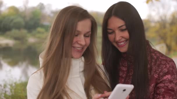 Dos hermosas chicas están caminando en el parque de otoño, viendo una foto en el teléfono — Vídeos de Stock