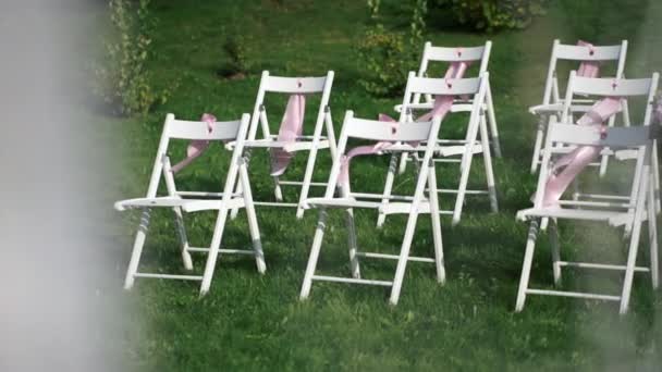 Weiße Stühle mit rosa Schleifen, die im Wind wehen. Hochzeitsdekor für die Registrierung unter freiem Himmel. — Stockvideo