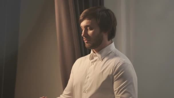 Brutal handsome man dressed in wedding day. The groom goes to the window, she puts on a red tie on a close-up of neck. The camera moves to the left. The frame is filled with light. The Big Day. — Stock Video