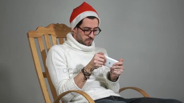 Amazing man in glasses and a red cap of Santa Claus playing a game on your phone — Stock Video