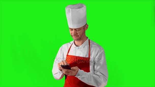 Bonito chef adulto digitando no smartphone, então ele olhando para a câmera e sorrindo em um fundo cinza — Vídeo de Stock