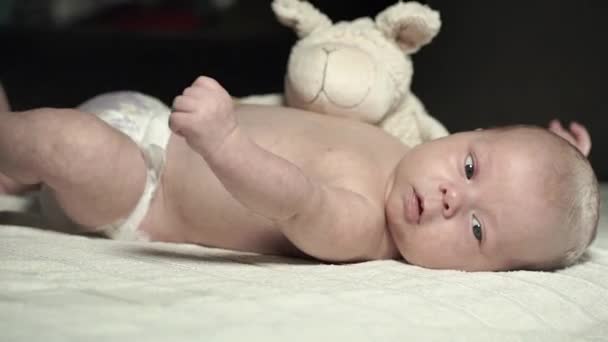 Een charmante baby ligt op een bed met een knuffel en actief beweegt zijn armen — Stockvideo