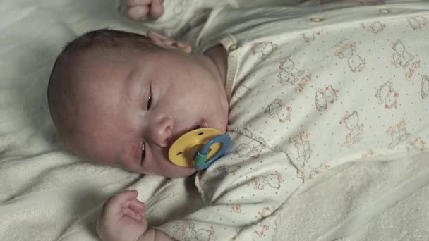 Een charmante baby met fopspeen in zijn mond ligt op een bed. Het licht verdwijnt langzaam — Stockvideo