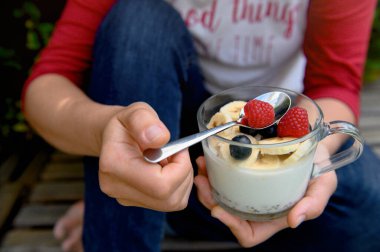 Sağlıklı bir kahvaltı yapıyorum. Yoğurt, granola, chia tohumu, taze ve kuru meyve, muz, yabanmersini, bir kadının elinde şeffaf bir bardakta ahududu. Temiz yemek, diyet, detoks, vejetaryen yemek.