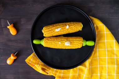Haşlanmış, olgun mısır ve tereyağı, ahşap bir masada, üzerinde sarı peçete olan siyah bir seramik tabakta. Düz yatıyordu.