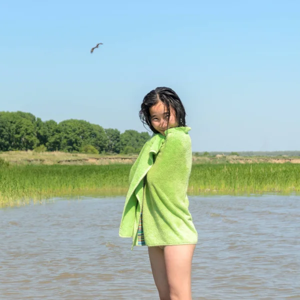 Beautiful Asian girl smiling is wrapped in a green towel with wet hair in swimsuit on the background of the birds the eagle and the trees of the forest to the cliff and grass near the water. — Stock Photo, Image