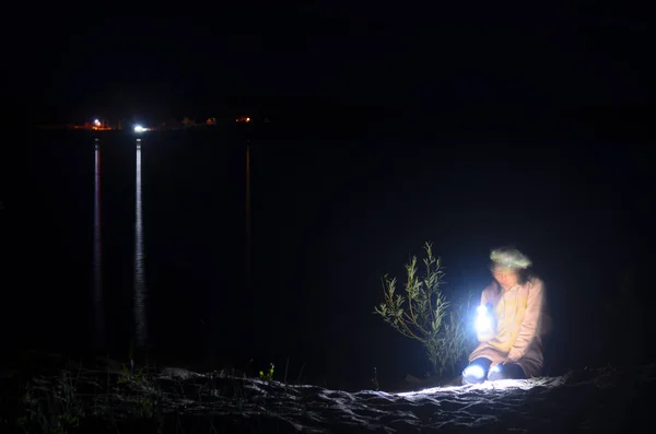Verschwommene Silhouette eines Mädchens in Jacke und Mütze, auf den Knien sitzend, mit einer Taschenlampe in der Nacht von den Blättern des Busches am Ufer des dunklen Flusses mit einem roten Lichtreflex der Lampe. — Stockfoto