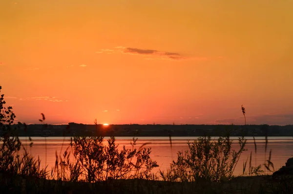 Součástí jasném slunečním disku na oranžové západy slunce na obzoru na obloze nad řekou trávy nad domy vesnice v Rusku. — Stock fotografie