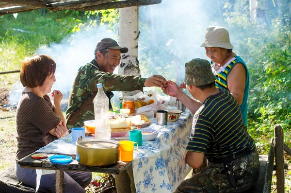 A joyful family feast in the Northern Yakut forest in the afternoon behind the smoke of a fire with elderly Asian women and men passing food.